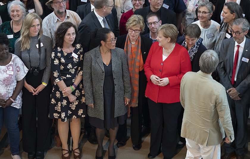 Dr. Angela Merkel steht mit mehreren zurückgekehrten Fachkräfte in einer Menschengruppe zusammen.