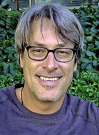 Porträt eines Mannes mit Brille, der draußen vor einer grünen Hecke lächelt.