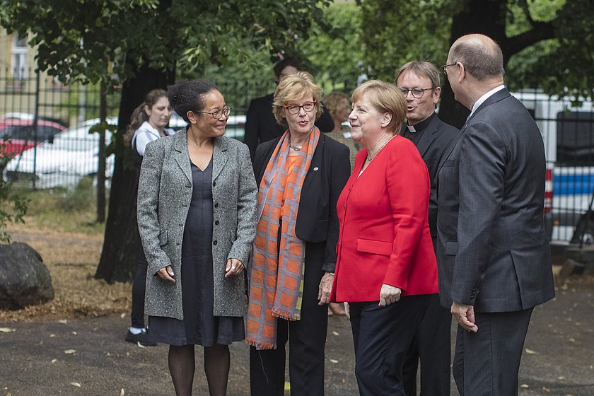 Fünf Menschen nebeneinander und unterhalten sich. Sie sind förmlich gekleidet. In der Mitt steht Angela Merkel in einem roten Blazer. 