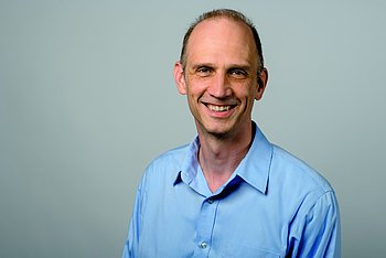 Portrait von Oliver Knabe. Ein lächelnder Mann mit kurzen Haaren, der ein hellblaues Hemd trägt. Er blickt freundlich in die Kamera vor einem neutralen grauen Hintergrund