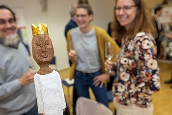 Im Vordergrund steht eine kunstvolle Figur, die einen goldenen Kopf mit einer glänzenden Krone hat. Der Kopf ist detailliert gestaltet und erinnert an eine Skulptur, während der Körper in weißem Gewand dargestellt ist. Im Hintergrund stehen lachend mehrere Personen, die die festliche Atmosphäre unterstreichen.