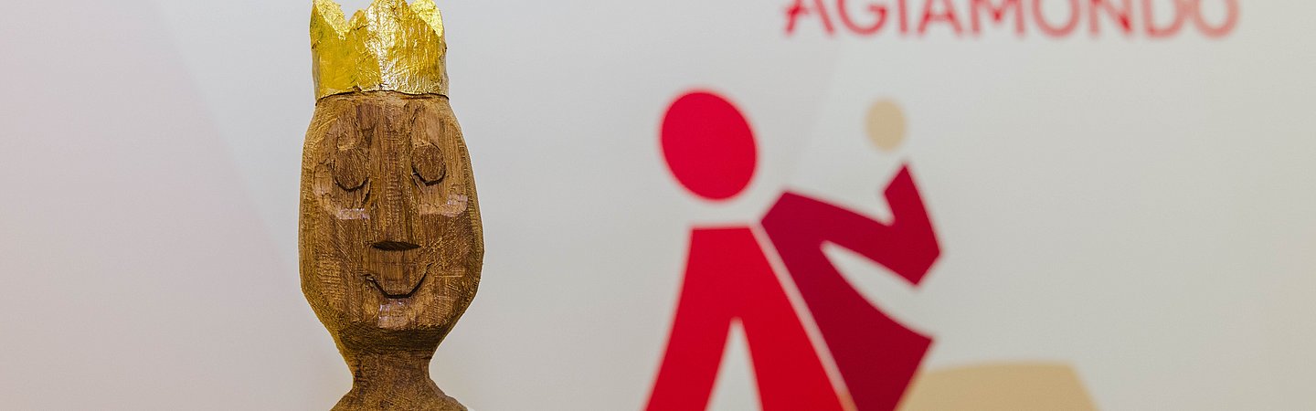 Die kunstvolle Figur des Engagementpreises von AGIAMONDO, mit einem goldenen Kopf und einer glänzenden Krone, sowie einem Körper in weißem Gewand. Im Hintergrund ist der Schriftzug 'Engagementpreis AGIAMONDO' und ein rotes Logo mit einer stilisierten Figur zu sehen.