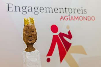 Die kunstvolle Figur des Engagementpreises von AGIAMONDO, mit einem goldenen Kopf und einer glänzenden Krone, sowie einem Körper in weißem Gewand. Im Hintergrund ist der Schriftzug 'Engagementpreis AGIAMONDO' und ein rotes Logo mit einer stilisierten Figur zu sehen.