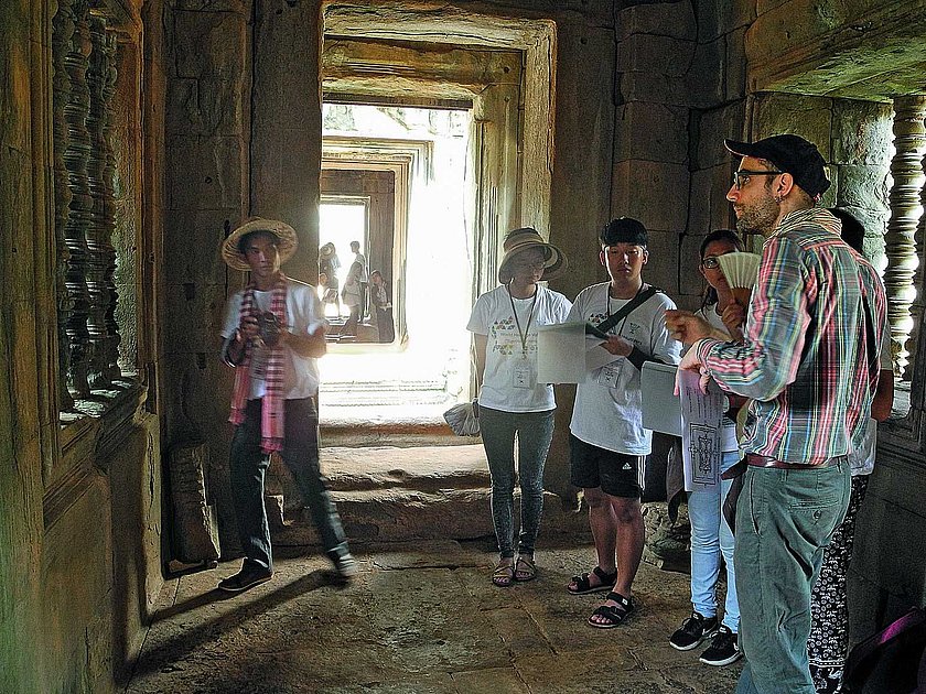 Eine Gruppe von Menschen erkundet das Innere einer historischen Ruine oder eines alten Gebäudes. Die Personen stehen in einem Korridor mit steinernen Wänden, und Licht fällt durch die Öffnungen im Hintergrund.