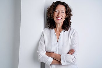 Portrait einer Frau mit schulterlangen braunen Locken und einem weißen Hemd. 