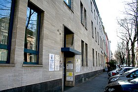 Das Bürogebäude der AGDD, ein mehrstöckiges Gebäude mit heller Fassade und großen Fenstern. Vor dem Eingang befinden sich Hinweisschilder, und entlang der Straße sind parkende Autos und Bäume zu sehen.