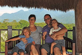 Eine Familie sitzt auf einer Veranda und blickt in die Kamera, mit einer malerischen Landschaft im Hintergrund.