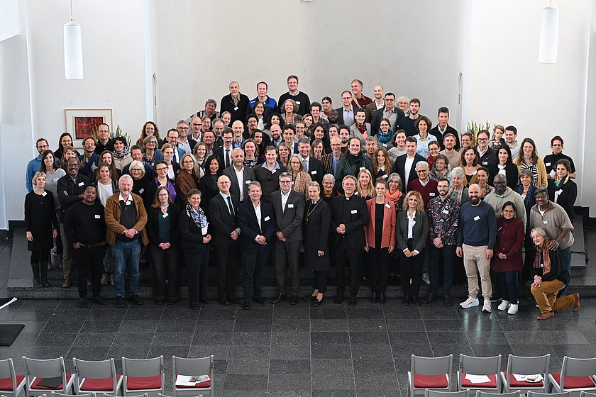 Ein großes Gruppenfoto, aufgenommen in einer Aula oder einem Konferenzraum, mit mehreren Reihen von Personen.