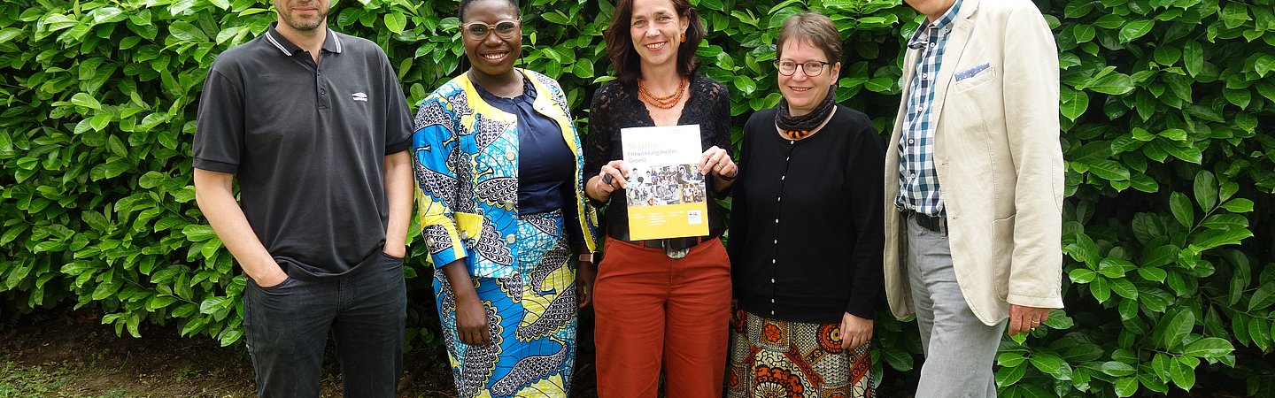 Fünf Personen stehen vor einer dichten, grünen Hecke und blicken lächelnd in die Kamera. Eine Frau in der Mitte hält ein Dokument oder ein Buch in den Händen. Die Gruppe trägt eine Mischung aus formeller und traditioneller Kleidung, die die Vielfalt und den Anlass hervorheben.