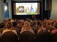 Eine große Gruppe von Menschen sitzt in einem abgedunkelten Raum und schaut auf eine Leinwand, auf der eine Präsentation läuft.