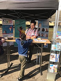 Ein dunkelhaariger Mann mit Bart und Brille im bunten Hemd steht an einem Stehtisch. Im Hintergrund befinden sich Rollups, auf denen "Vorlesen macht stark" und "Macht zusammen Bielefeld" steht. Ein Junge auf der anderen Seite des Tisches spricht mit dem Mann. 