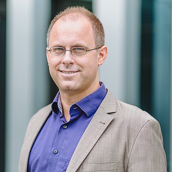 Ein Profil von einem Mann ist zu sehen. Er trägt ein lilanes Hemd und einen grauen Blazer.