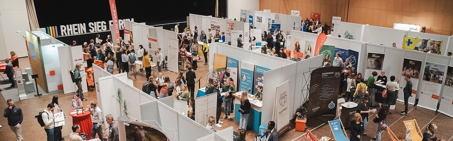 Eine große Messe mit verschiedenen Ständen und Besuchern in einer hellen und geräumigen Halle. Menschen erkunden die Ausstellungsstände.