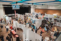 Eine große Messe mit verschiedenen Ständen und Besuchern in einer hellen und geräumigen Halle. Menschen erkunden die Ausstellungsstände.