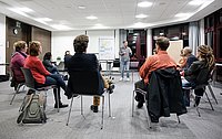 Menschen sitzen im Kreis auf Stühlen und diskutieren in einer offenen und entspannten Atmosphäre. Eine Flipchart steht im Hintergrund.