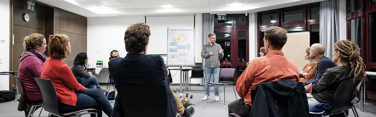 Menschen sitzen im Kreis auf Stühlen und diskutieren in einer offenen und entspannten Atmosphäre. Eine Flipchart steht im Hintergrund.