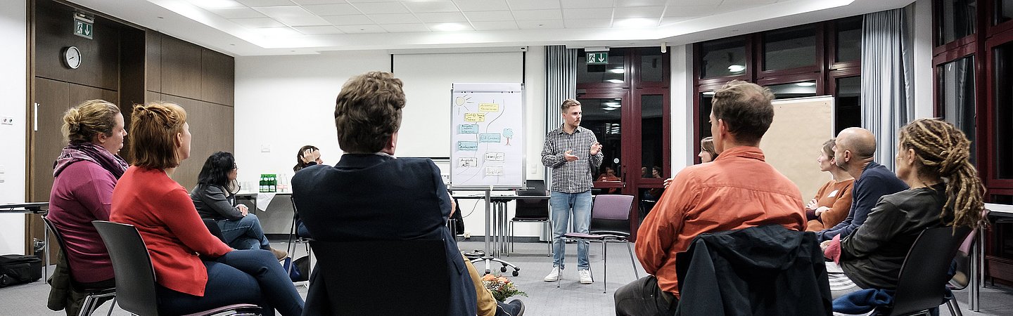 Menschen sitzen im Kreis auf Stühlen und diskutieren in einer offenen und entspannten Atmosphäre. Eine Flipchart steht im Hintergrund.