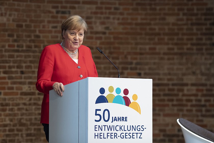 Angela Merkel in einem roten Blazer steht an einem weißen Rednerpult und hält eine Ansprache. Auf dem Pult ist das Logo mit der Aufschrift '50 Jahre Entwicklungsgesetz' und farbigen Symbolfiguren abgebildet. Der Hintergrund besteht aus einer strukturierten Ziegelwand.