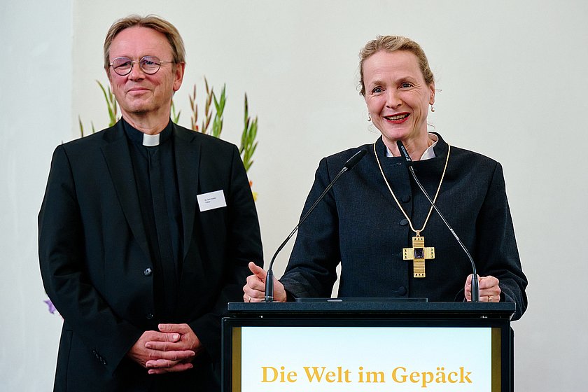 Zwei Personen, ein Mann und eine Frau, stehen nebeneinander auf einer Bühne und lächeln, während sie sprechen. Im Hintergrund ist ein Blumengesteck sichtbar.