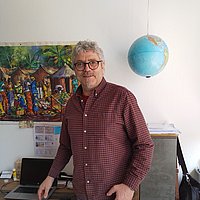 Ein Mann mittleren Alters mit grauen Haaren steht in einem Büro. Hinter ihm ist ein Globus sowie eine bunte Karte an der Wand zu sehen. Er trägt ein bordeauxfarbenes Hemd und blickt freundlich in die Kamera.