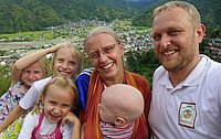 Eine Familie mit sechs Personen, bestehend aus zwei Erwachsenen, Mann und Frau und vier Kindern, lächelt in die Kamera. Im Hintergrund ist eine grüne Hügellandschaft mit vereinzelten Häusern zu sehen.