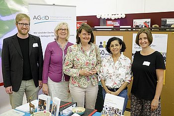 Fünf Personen stehen nebeneinander und schauen in die Kamera. Im Hintergrund der Stand der AGdD.