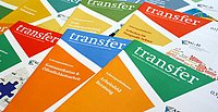 Mehrere bunte Broschüren mit der Aufschrift 'Transfer', gestapelt auf einem Tisch.