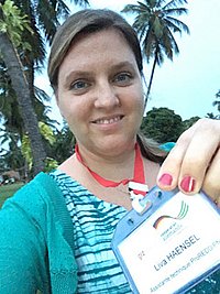  Eine Frau hält ein Namensschild in die Kamera und lächelt. Sie trägt ein blau-grünes Oberteil und eine rote Kordel um den Hals. Daran hängt ein Schild mit dem Namen Liva Hänsel. Im Hintergrund sind Palmen und eine grüne Wiese zu sehen.
