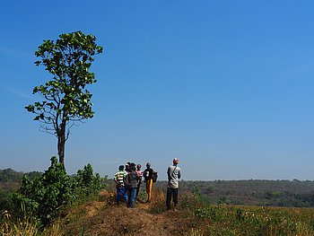 Mehrere Menschen stehen auf einem offenen Feld und führen ein Interview durch, mit einem klaren blauen Himmel im Hintergrund.