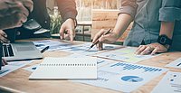 Mehrere Personen arbeiten gemeinsam an einem Tisch, der mit Papieren, Diagrammen und einem Notizbuch bedeckt ist. Hände halten Stifte und zeigen auf verschiedene Stellen auf den Dokumenten. Die Szene vermittelt eine Atmosphäre von Teamarbeit und Planung.
