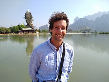 Felix steht vor einem Fluss in Myanmar mit einer malerischen Landschaft und Bergen im Hintergrund.