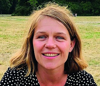 Porträt einer lächelnden Frau vor einer natürlichen Kulisse mit Wiese und Bäumen im Hintergrund.