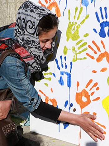 Eine Frau vor einer Leinwand kniend auf der Handabdrücke in bunten Farben zu sehen sind. 