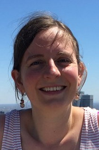 Porträt einer Frau mit kurzen Haaren, die im Freien lächelt, vor einem klaren blauen Himmel.