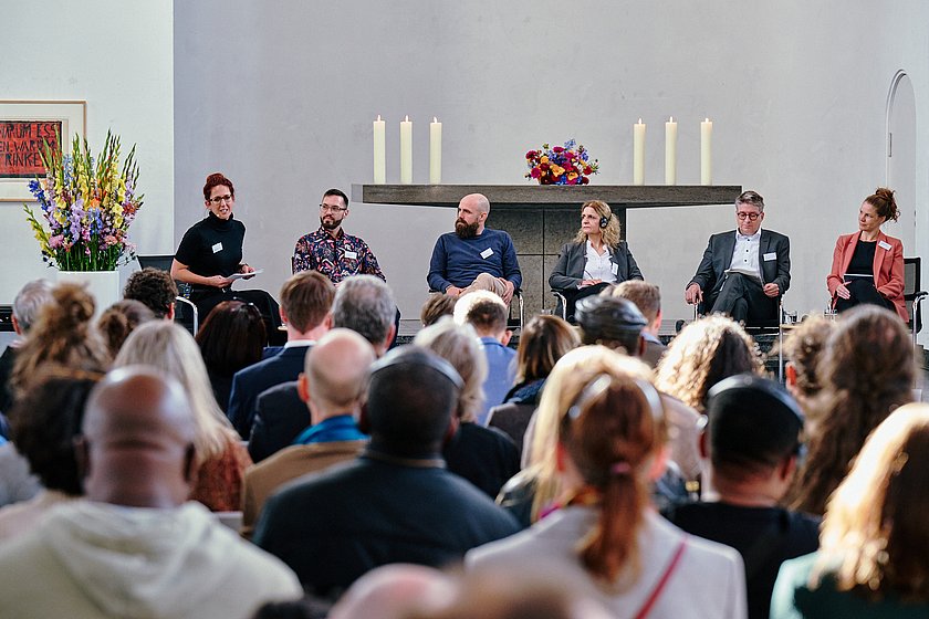 Ein Blick über die Köpfe eines Publikums hinweg auf eine Bühne, auf der mehrere Personen sitzen, umgeben von Kerzen und Dekoration.