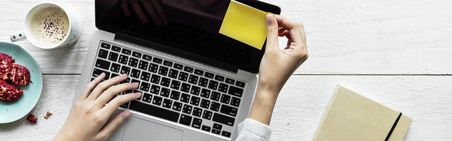 Eine Person arbeitet an einem Laptop mit einer Tasse Kaffee und einem Notizbuch daneben, auf einer weißen Tischoberfläche.