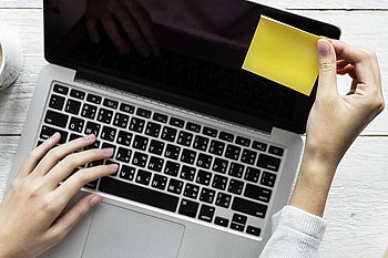 Eine Person arbeitet an einem Laptop mit einer Tasse Kaffee und einem Notizbuch daneben, auf einer weißen Tischoberfläche.