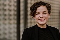Porträtfoto einer Frau mit lockigem Haar und schwarzer Kleidung, aufgenommen vor einem neutralen Hintergrund.