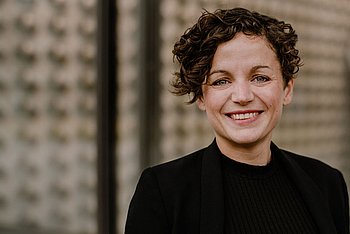 Porträtfoto einer Frau mit lockigem Haar und schwarzer Kleidung, aufgenommen vor einem neutralen Hintergrund.
