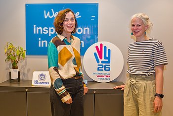 Zwei Frauen stehen nebeneinander. Zwischen ihnen das International Year of Volunteers 2026-Logo. 