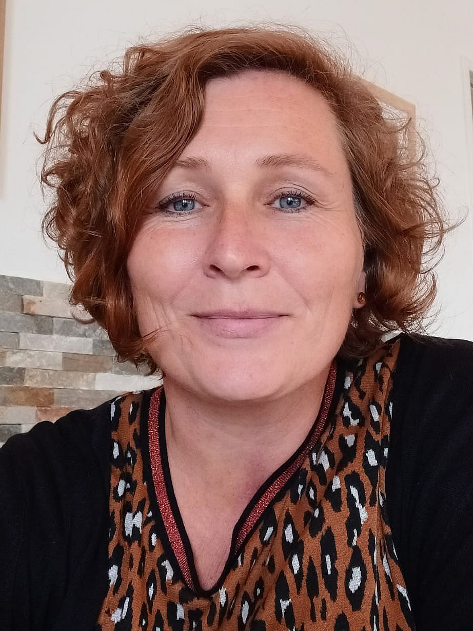 Eine Frau mittleren Alters mit kurzen lockigen Haaren in schwarzer Strickjacke lächelt in die Kamera. 
