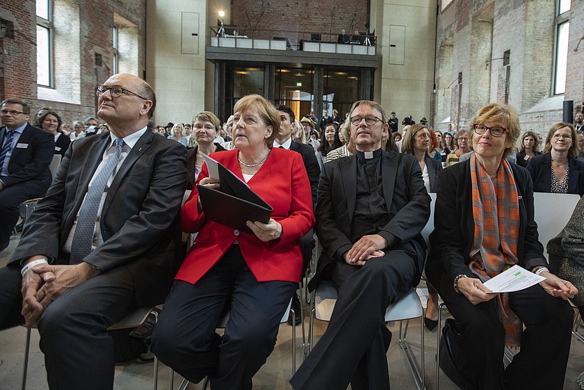 Vier Personen sitzen in der ersten Reihe einer Veranstaltung in einem großen Saal mit hohen Decken und Ziegelwänden. Die Person in der Mitte trägt einen roten Blazer und hält ein Dokument in der Hand. Im Hintergrund sind weitere sitzende Teilnehmer und die Bühne erkennbar.