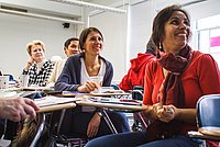 Eine Gruppe von Menschen sitzt in einem Seminarraum und hört aufmerksam einem Vortrag zu, mit einem Fokus auf eine Frau in einem roten Oberteil, die lächelnd nach vorne schaut.