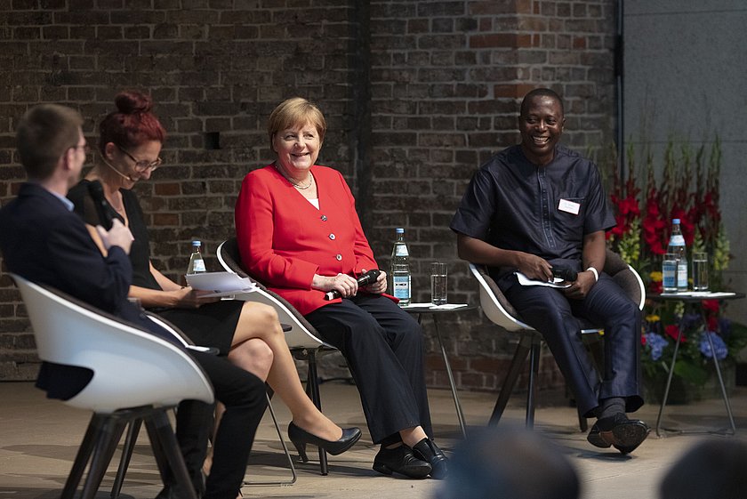 Eine Podiumsdiskussion. Vier Personen sitzen in einem Halbkreis auf Stühlen. In der Mitt sitzt Angela Merkel in einem roten Blase rund führt das Gespräch. 