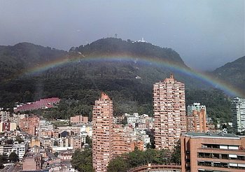 Eine Stadtansicht mit hohen Wohngebäuden im Vordergrund, umgeben von grünen Hügeln. Ein Regenbogen spannt sich über die Landschaft, während der Himmel wolkig ist.