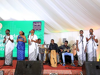 Eine Musikgruppe auf der Bühne. Die Sängerinnen in traditioneller Kleidung performen vor einem Publikum, begleitet von Musikern im Hintergrund. Die Bühne ist mit einem grünen Banner dekoriert und befindet sich unter einem Zelt.