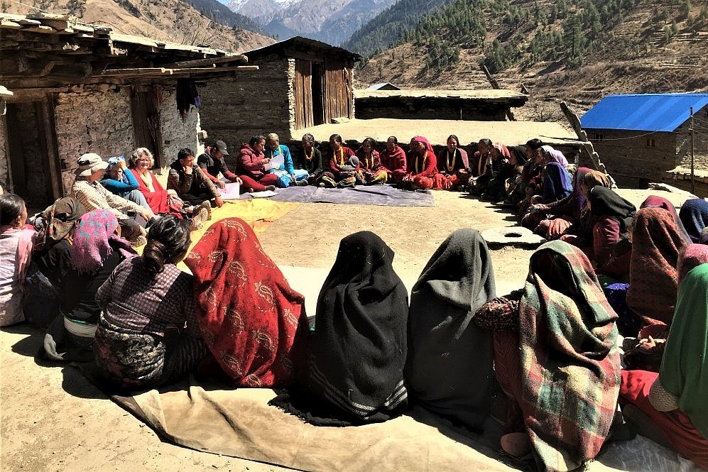 Eine Gruppe von Frauen sitzt im Freien in einem großen Kreis, viele von ihnen in farbenfrohen traditionellen Gewändern. Im Hintergrund sind einfache Steinhäuser und eine malerische Berglandschaft mit schneebedeckten Gipfeln zu sehen. Die Szene vermittelt eine gemeinschaftliche Atmosphäre in einer abgelegenen ländlichen Umgebung.
