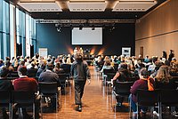 Ein Konferenzraum mit vielen Sitzreihen, in denen Teilnehmer einer Präsentation folgen. Ein großes Display ist im Hintergrund sichtbar.