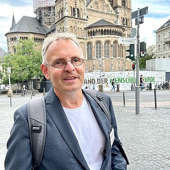Ein Mann mit Brille und Rucksack steht auf einem öffentlichen Platz vor einer historischen Kirche. Im Hintergrund sind Bäume und eine kleine Menschenansammlung zu sehen.