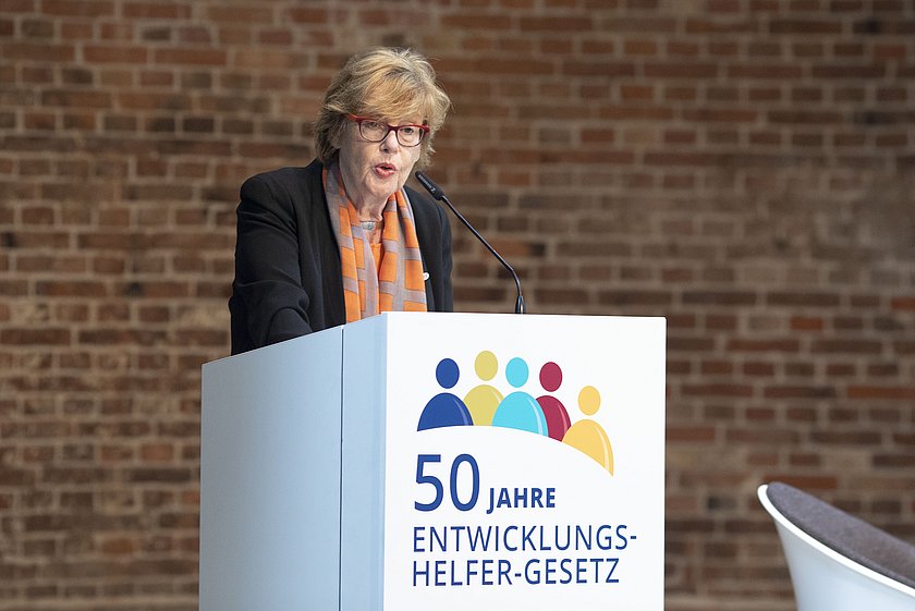 Dr. Cornelia Füllkrug-Weitzel Präsidentin von Brot für die Welt eröffnet die Veranstaltung „Die Welt im Gepäck“ am Rednerpult. Mit der Aufschrift '50 Jahre Entwicklungshelfer-Gesetz'. Die Frau trägt eine Brille, eine schwarze Jacke und ein orangefarbenes Halstuch. Im Hintergrund ist eine Backsteinwand zu sehen.
